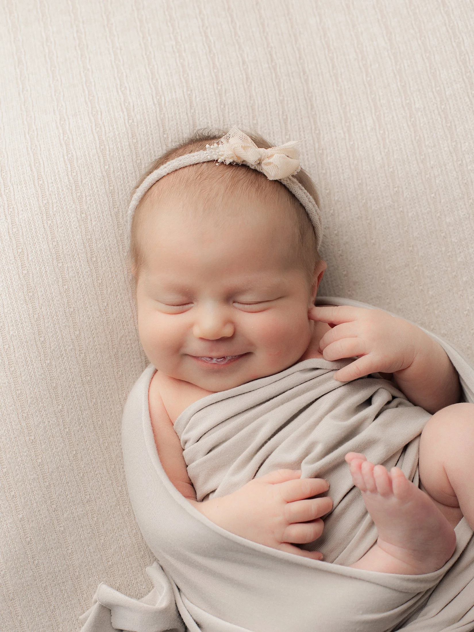 Hymyilevä vauva makaa pehmeällä alustalla, kapaloitu vaalean ruskeaan kankaaseen, hiuksissa pieni rusettipanta.