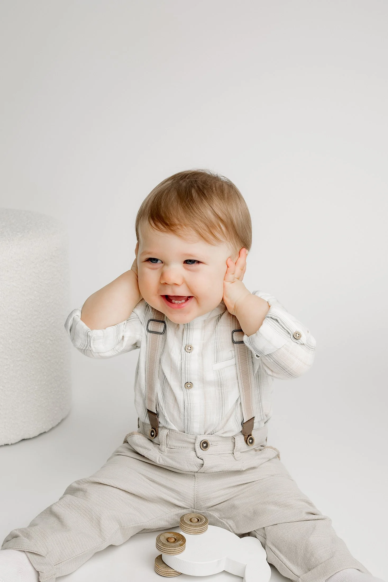 Pieni poika istuu lattialla, nauraa kädet korvillaan. Yllään vaalea asu, housuissa henkselit.