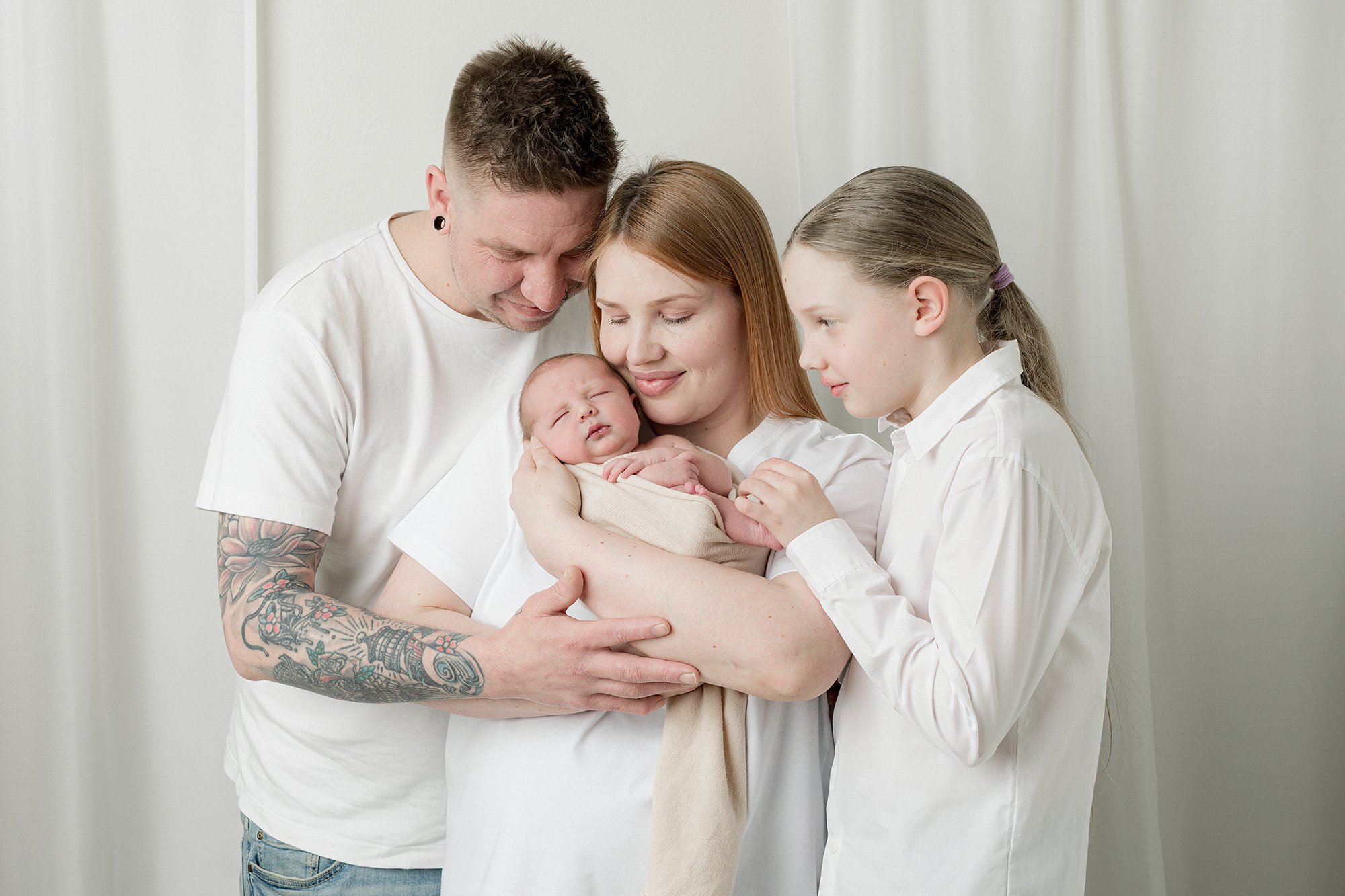 Perhe, jossa vanhemmat ja kaksi lasta, halaavat ja katsovat lasta, joka on vauva, sisällä seinän vieressä