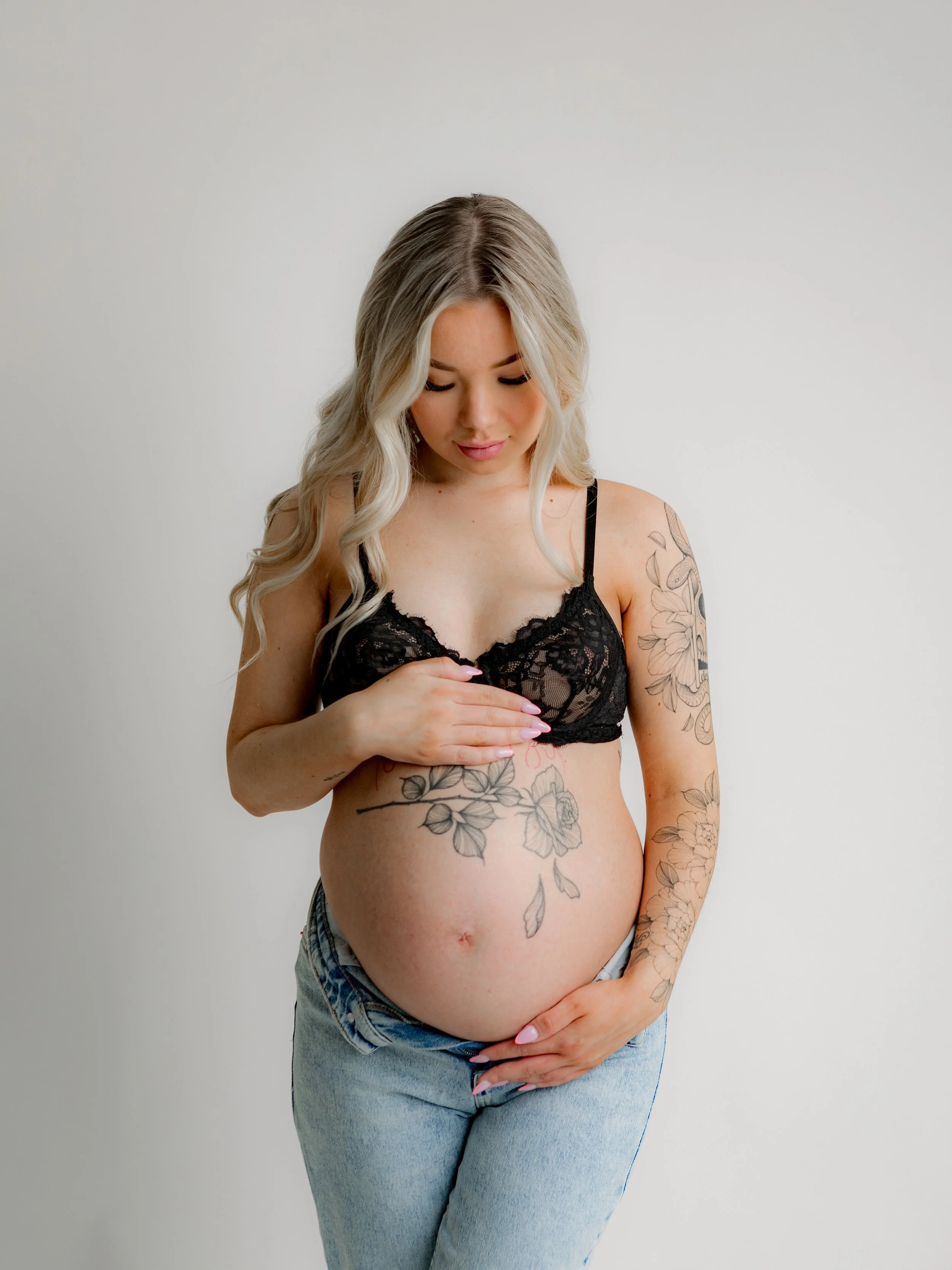 Raskaana oleva nainen, jossa on tatuointeja vatsassa ja käsivarsissa, katsoo alas, pitäen käsivarttaan vatsallaan, pukeutuneena mustaan pitsiliiviin ja farkkuhousuihin.