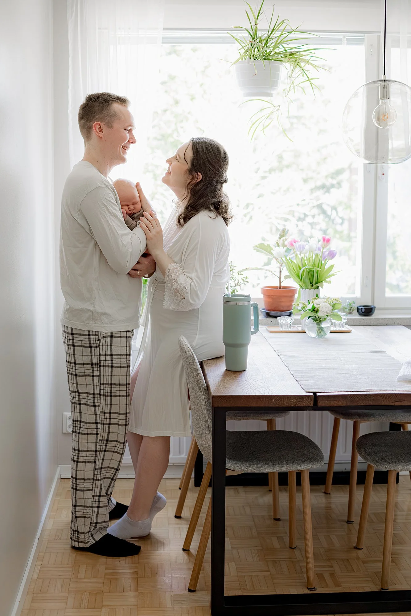 Äiti ja isä seisoo keittiössä ikkunan edessä sylissään pieni vastasyntynyt. Taustalla viherkasveja.