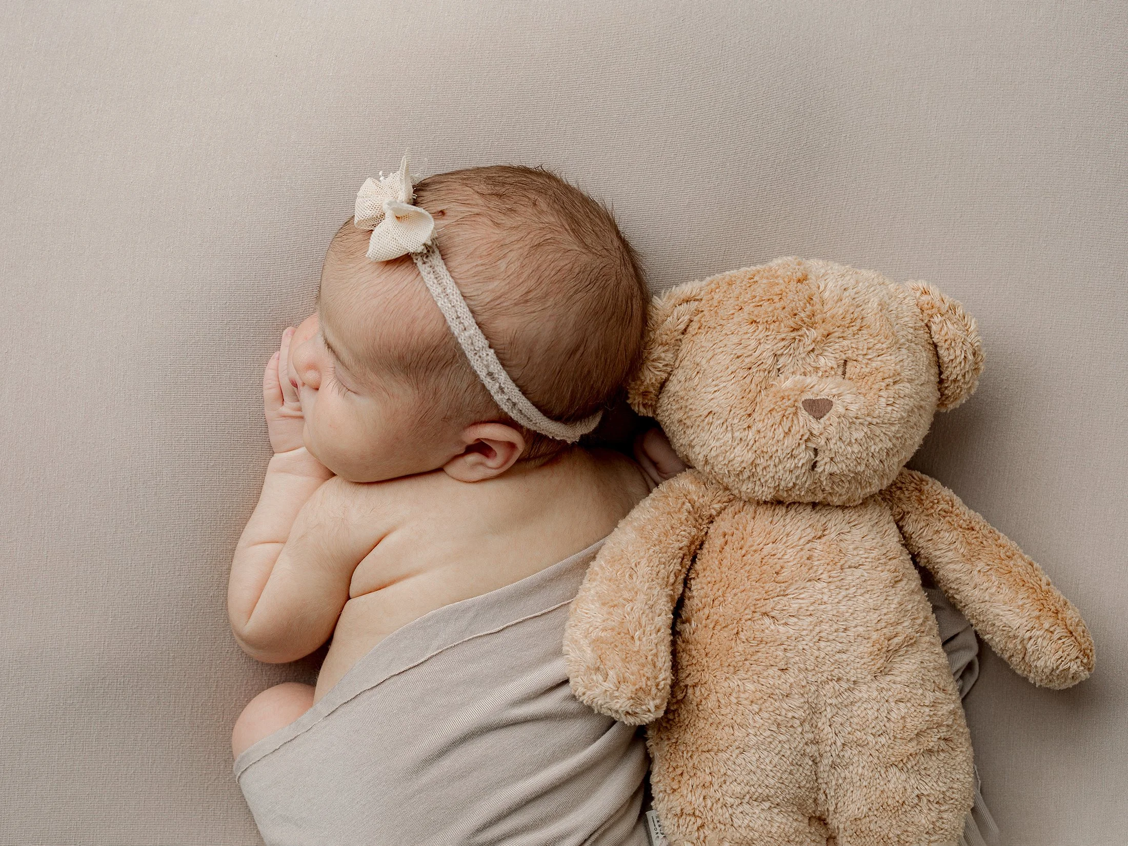 Vastasyntynyt tyttö nukkuu vatsallaan nalle vieressä. Beige peitto ja lakana.