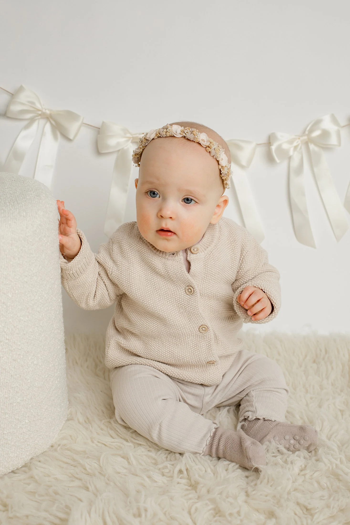 Pieni tyttö istuu vaalealla karvamatolla, taustalla rusetteja, yllään vaaleanruskea neule