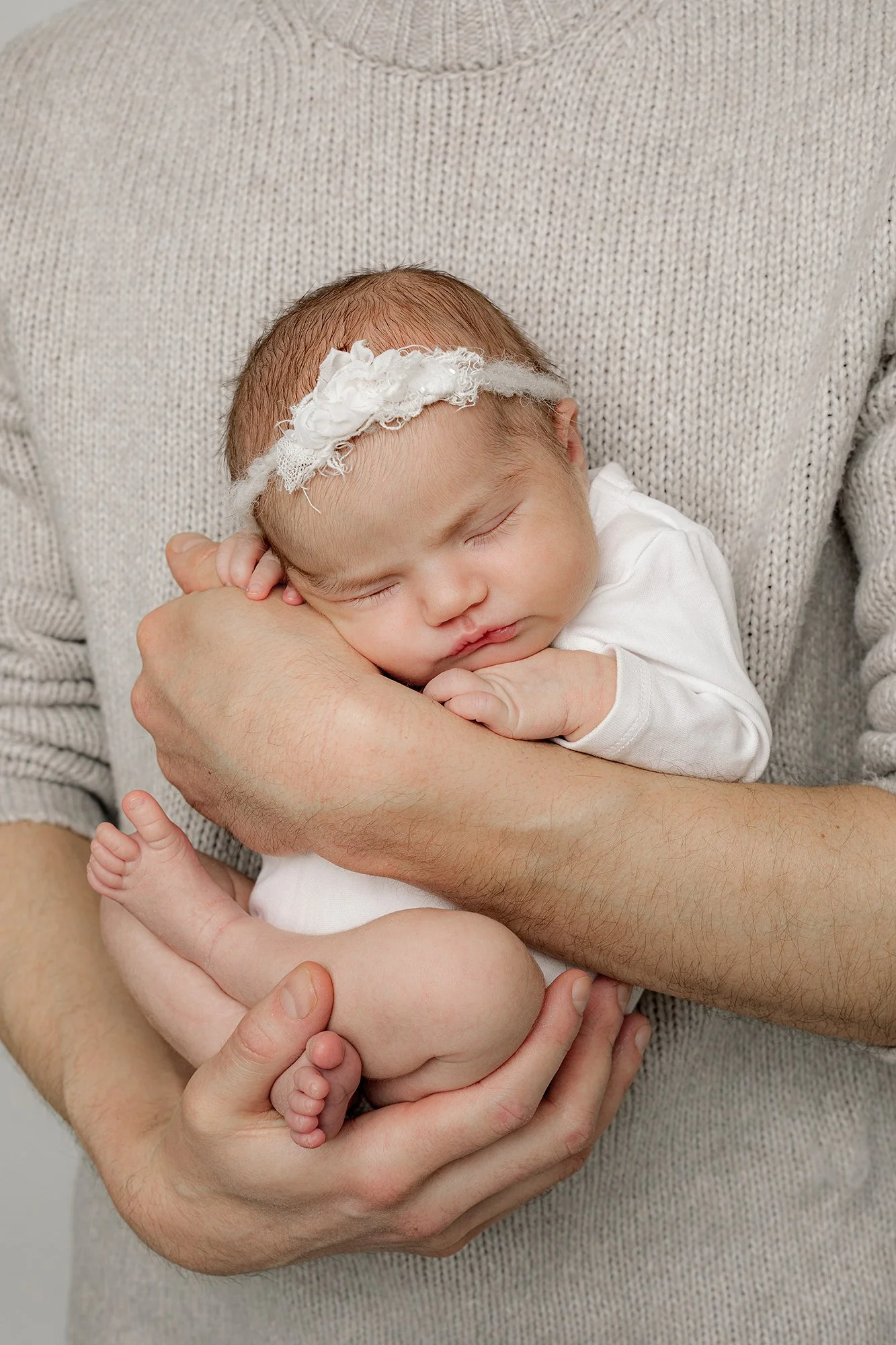 Vastasyntynyt tyttövauva isän sylissä pienellä kippuralla, yllään valkoinen body ja päässä valkoinen rusetti.