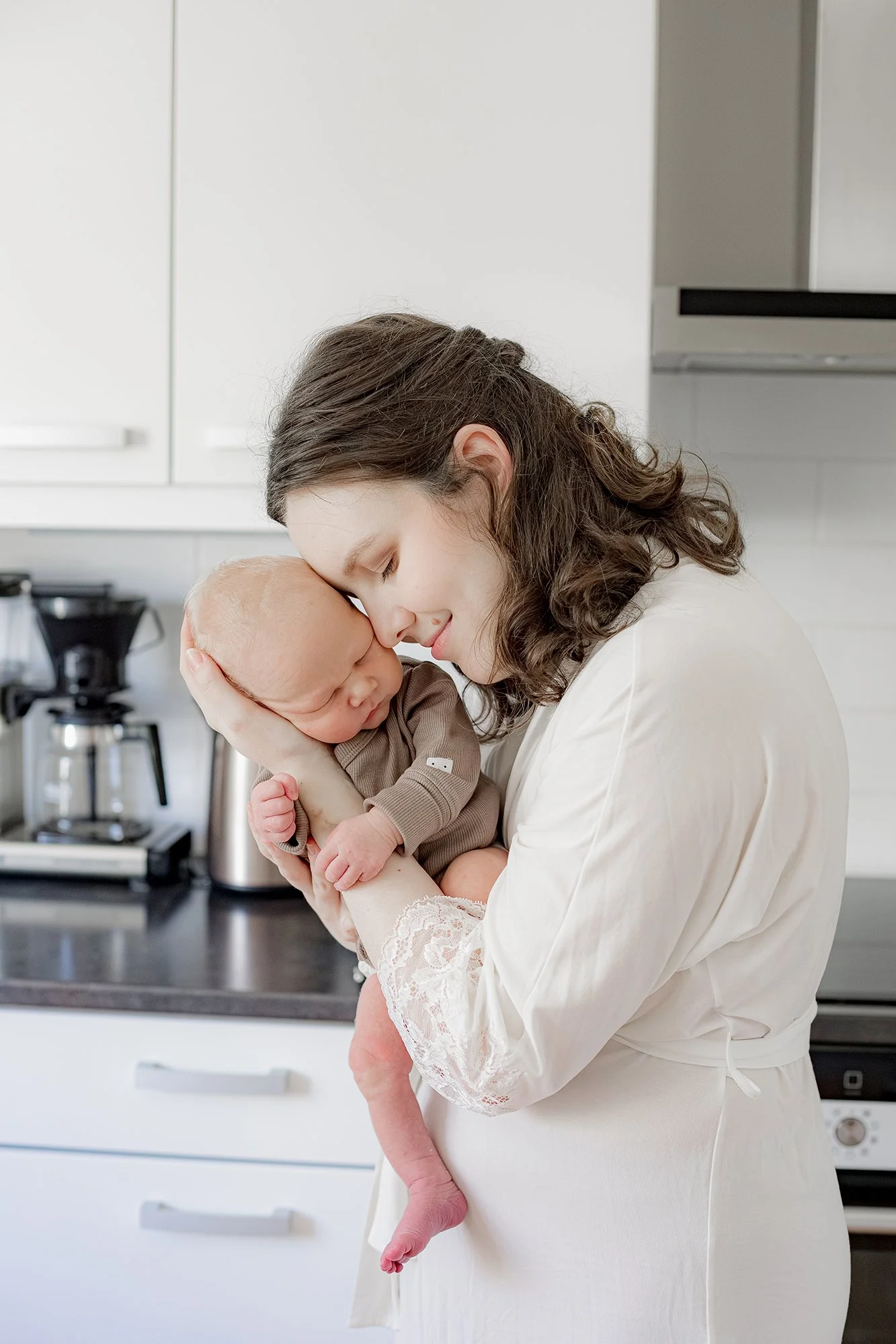 Äiti pitää hellästi käsivarsillaan vastasyntynyttä poikaansa. taustalla keittiö