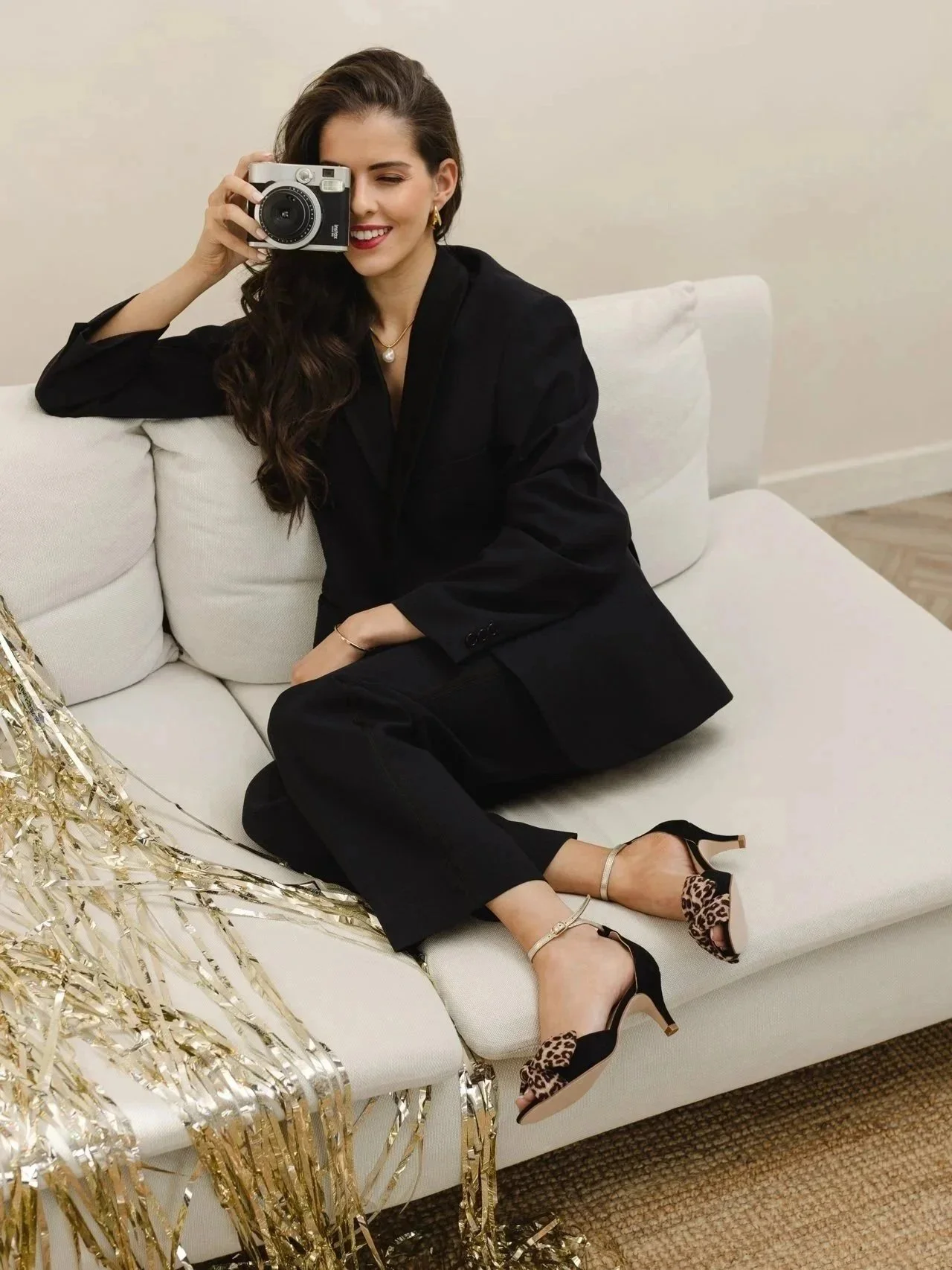 A woman sitting on a white sofa, taking a selfie with a camera, celebrating with gold tinsel decorations, wearing a black blazer, black pants, and leopard-print high heels.