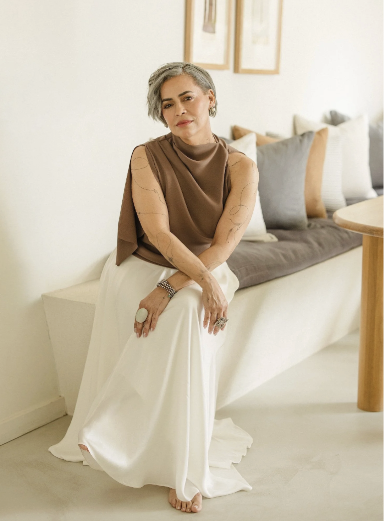 A woman with short gray hair, wearing a brown sleeveless top and white silk pants, sitting on a white bench or sofa with cushions in a bright room.