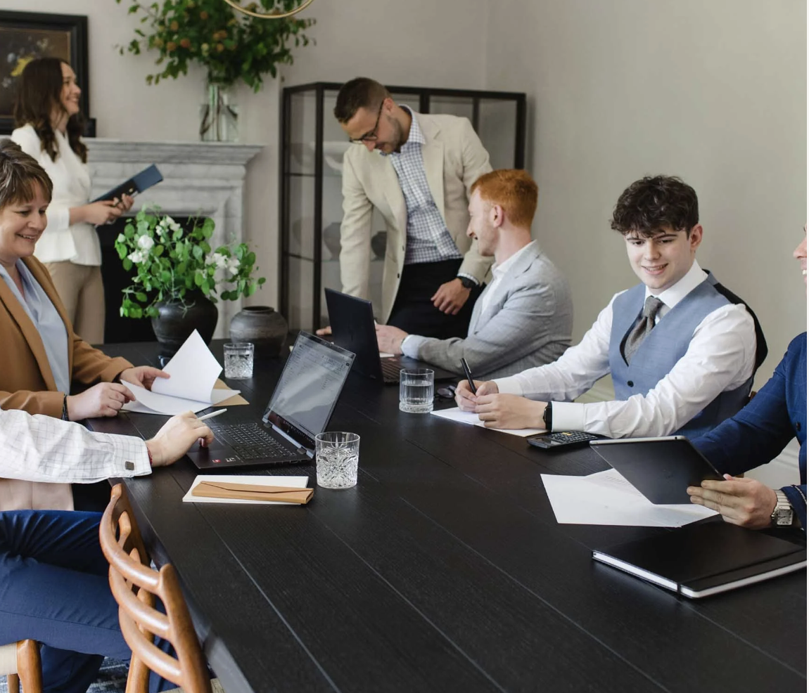 Business meeting with six people, five sitting at a table with laptops, tablets, and papers, one standing and smiling, in a well-lit modern conference room with plants and decorative items.