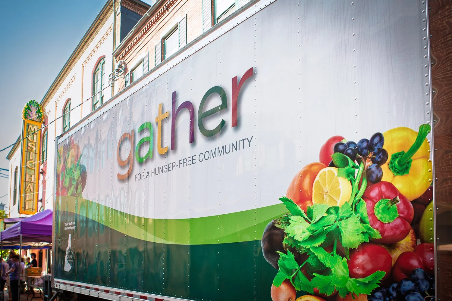 A community event banner promoting the homeless for a hunger-free community, with images of fruits including lemons, blueberries, red and yellow bell peppers, and greens.