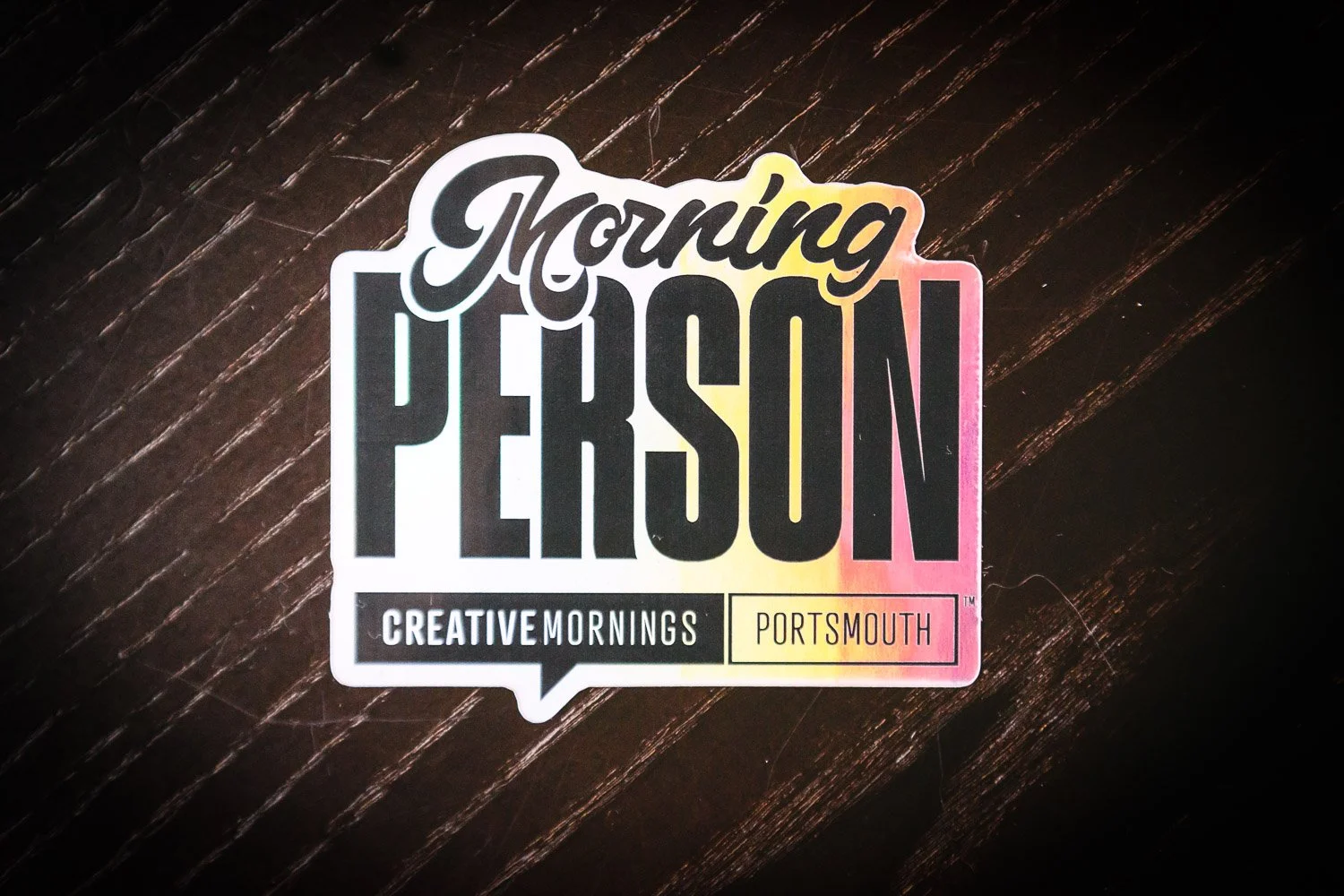 Sticker with text reading 'Morning Person' and smaller text 'Creative Mornings Portsmouth' on a dark wooden surface.