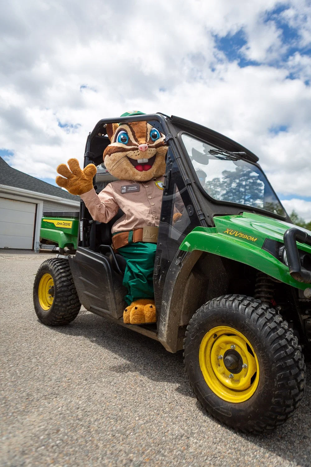 A person in a chipmunk mascot costume sitting in a green John Deere utility vehicle, waving. The scene is outdoors with a partly cloudy sky and a garage in the background.