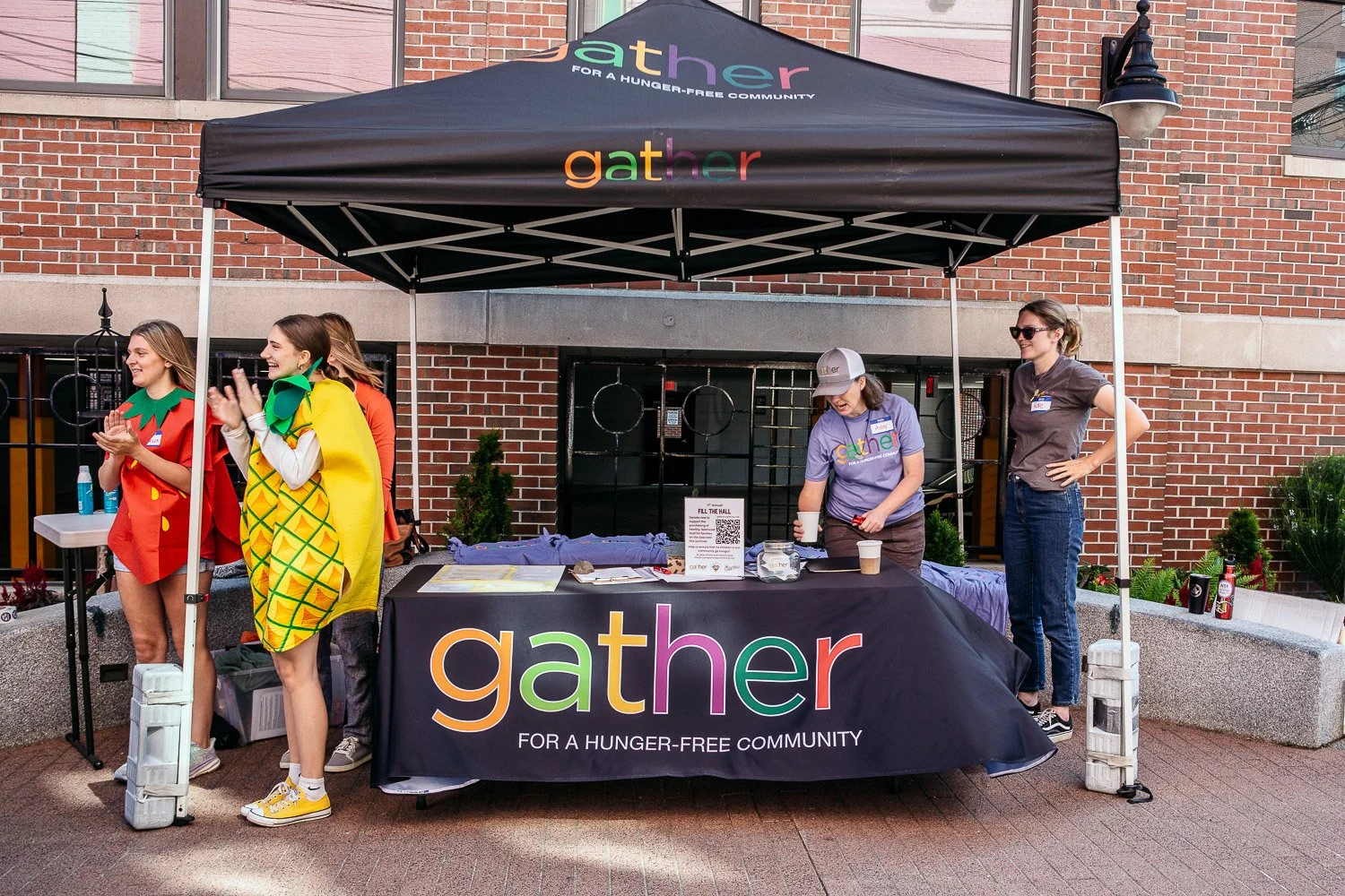 People at a community outreach booth under a black canopy with 'gather' written in colorful letters, promoting a hunger-free community. Four women, some dressed in colorful costumes, stand near the table with informational materials. One woman is pou
