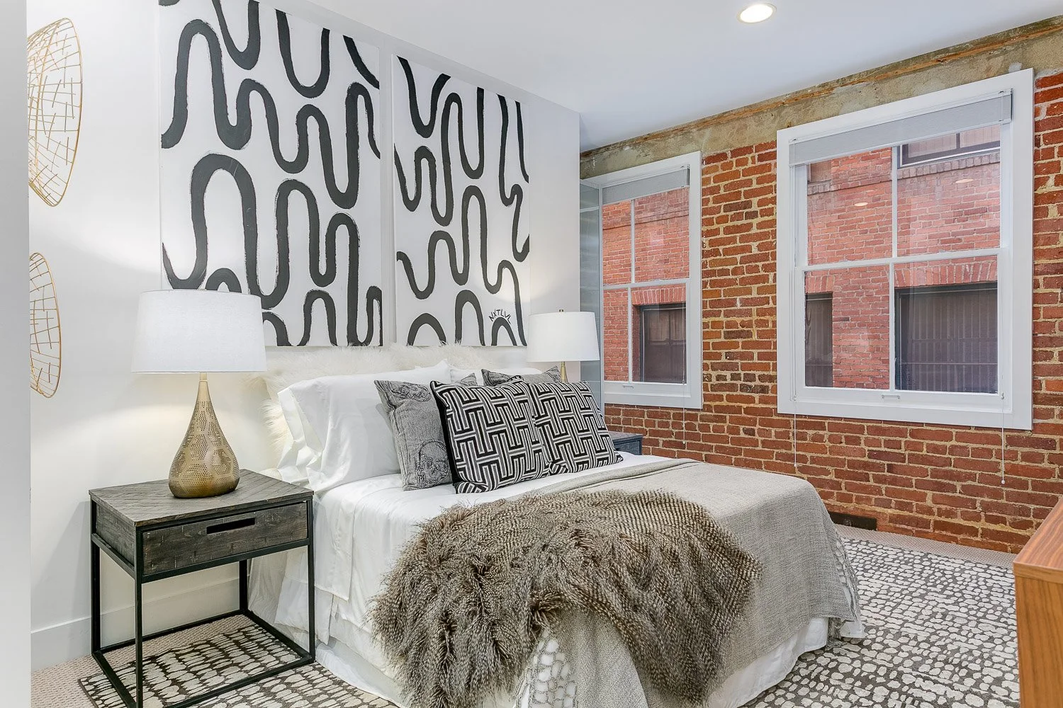 A bedroom with a bed featuring white and patterned pillows, a fur throw, and two white lamps on bedside tables, set against a wall decorated with a black abstract design and a brick wall with two large windows.