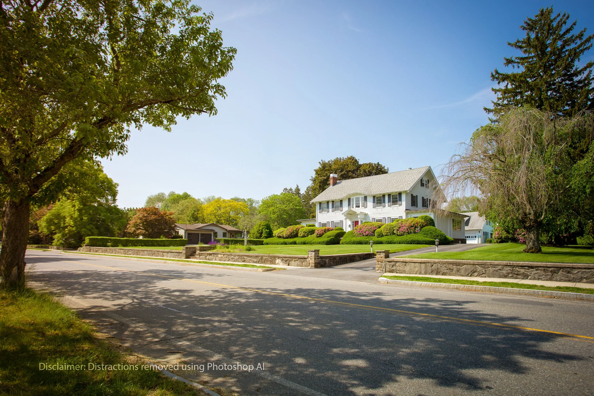 Large white house with a front porch, surrounded by well-manicured green lawns, bushes, and trees on a sunny day.