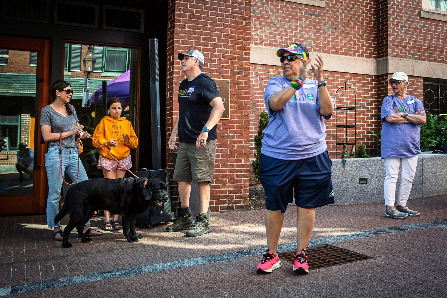 People gathered outside near a brick building, some wearing Gathering logo shirts, with a woman in colorful accessories and a man with a hat in the foreground. A woman on the left holds a leash with a black dog, while others are standing around, some