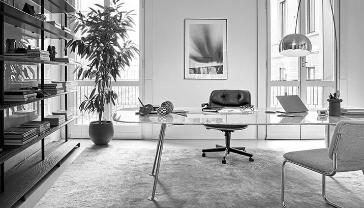 Modern home office with a glass desk, black leather chair, bookshelf, large window, potted plant, laptop, art print, and a minimalistic decor.