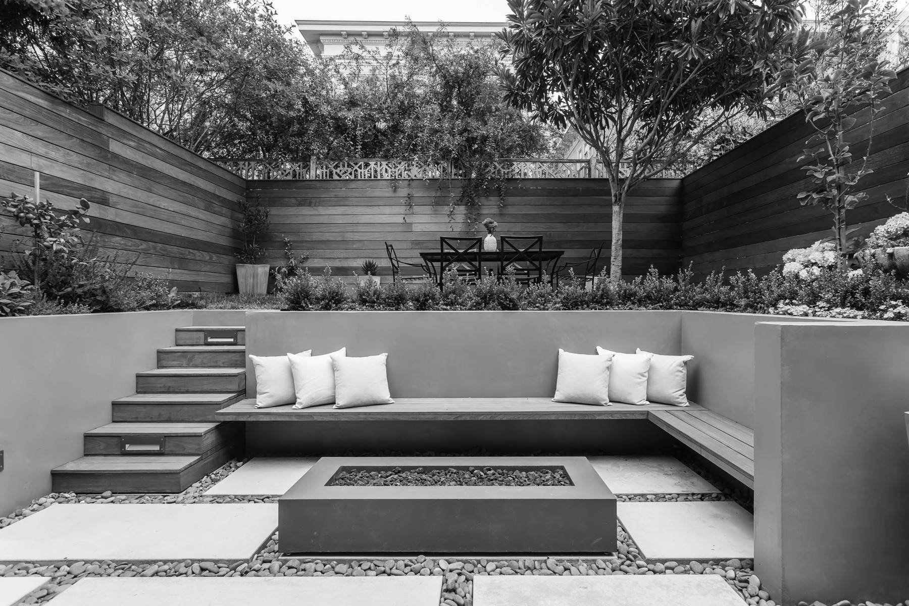 A black and white image of a backyard with a modern design. There are stairs leading to a built-in bench with several pillows. Directly in front is a rectangular fire pit surrounded by stone pavers and pebbles. In the background, there is a higher level with a patio table, chairs, and potted plants, enclosed by wooden fencing and lush trees.