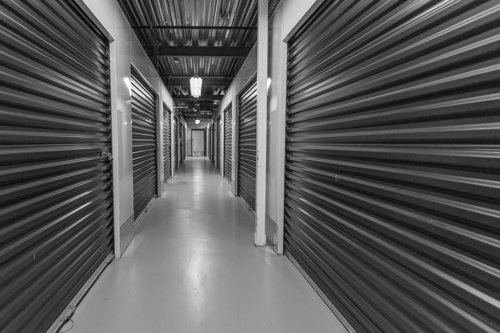 Black and white photo of a corridor with storage units, with metal rolling doors on each side and lights on the ceiling.