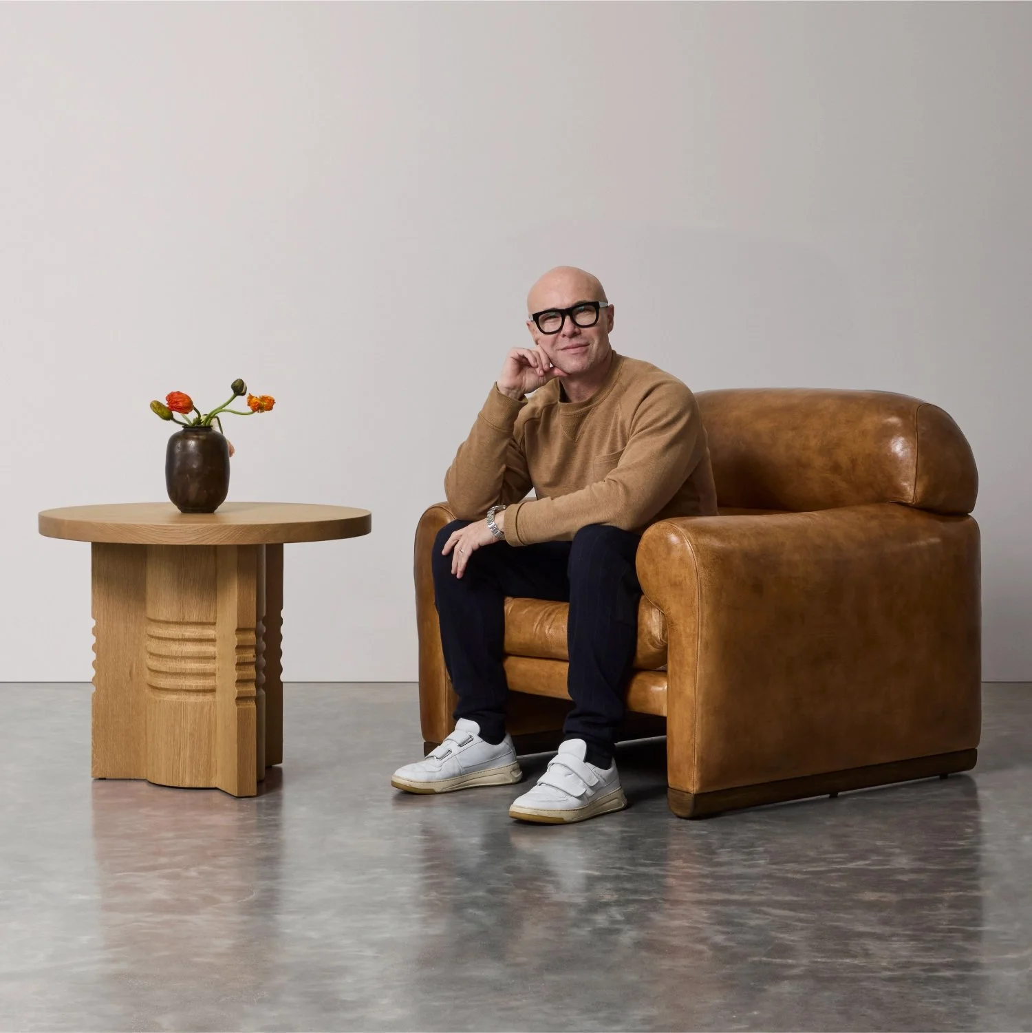 A man with glasses sitting on a brown leather armchair, wearing a tan sweater, black pants, and white sneakers. There is a small wooden table with a dark brown vase with orange flowers on the left, and a plain white wall in the background.