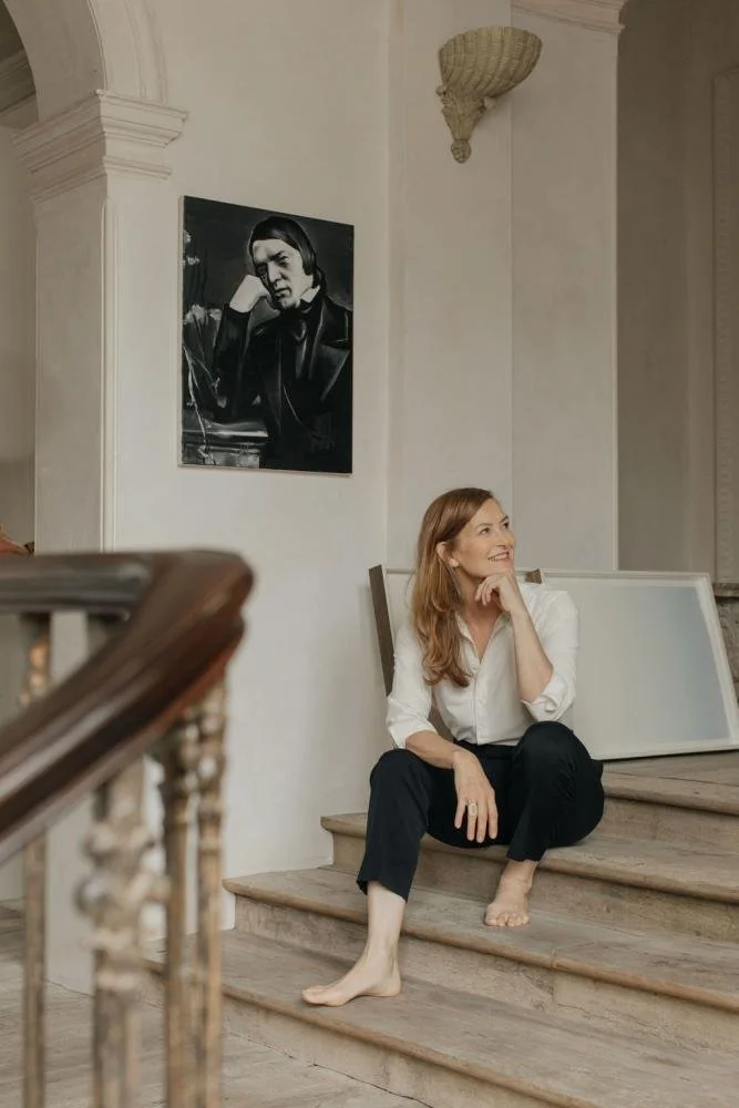 A woman with reddish-brown hair, wearing a white blouse and black pants, sits on the stairs barefoot, smiling and looking to her right in a well-lit interior space with white walls and a framed black-and-white photograph of a man wearing headphones hanging on the wall.