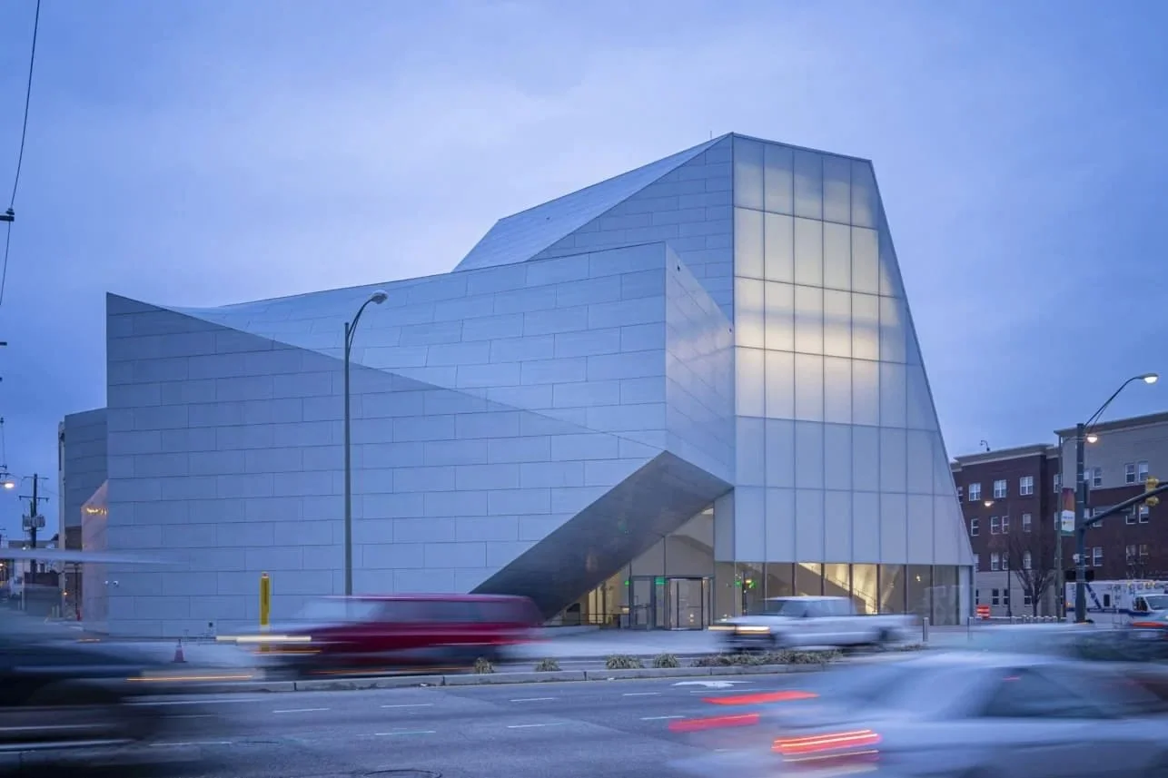 Modern building with angular design, glass and metal exterior, standing by a busy street with blurred cars passing by at dusk.