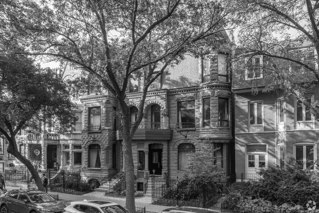 Black and white photo of a row of Victorian-style houses with prominent bay windows, decorative brickwork, and small front yards, lined with mature trees and parked cars.