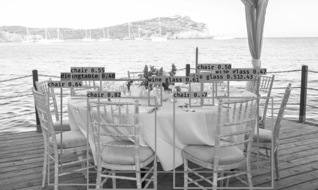 A table set for dining on a dock overlooking a lake with sailboats andland in the background; chairs, a wine glass, and utensils are on the table.