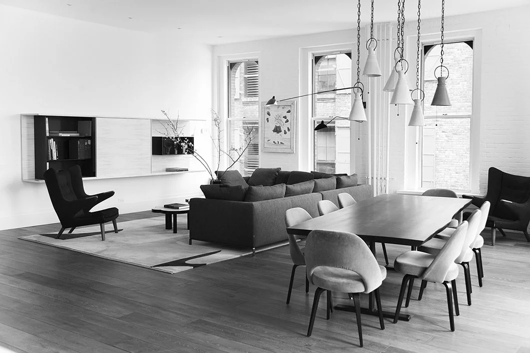 Modern living and dining room with a large gray sofa, black armchairs, a wooden dining table with eight chairs, pendant lighting, and large windows in a bright, minimalist space.