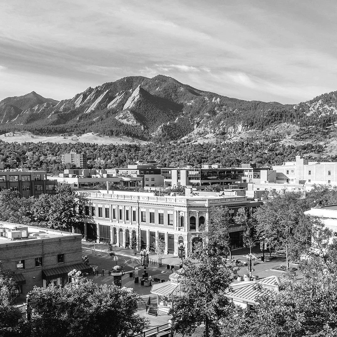 A black and white photo of a downtown city with old brick buildings, trees, and a mountain range in the background under a partly cloudy sky.