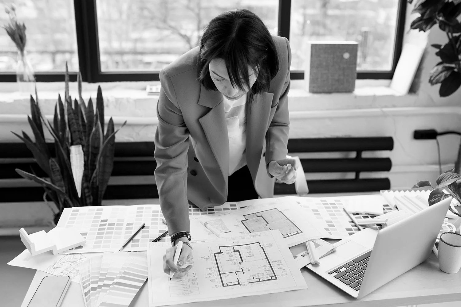 A woman in a blazer analyzing architectural blueprints on a desk, surrounded by color swatches, a laptop, and plants in a well-lit office meeting room.