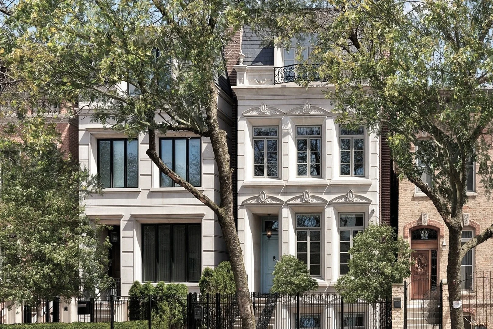 Front view of a white, multi-story residential building with decorative window moldings, surrounded by trees and a black iron fence.