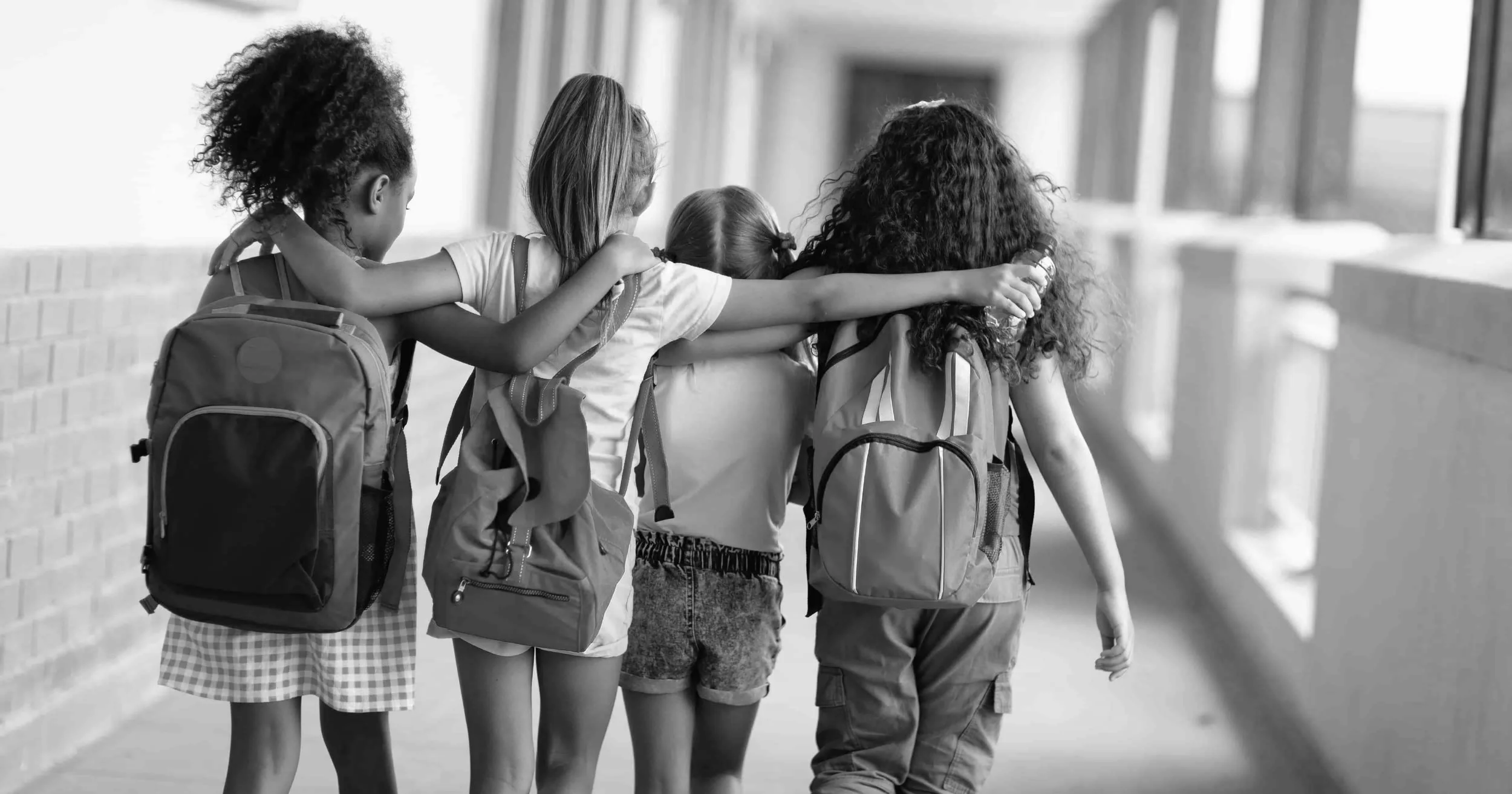 Four young girls with backpacks walking together in a school hallway, with their arms around each other in a friendly manner.