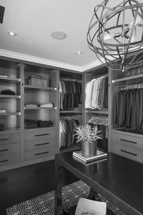 A walk-in closet with built-in shelving and hanging rods filled with clothes, a dark wooden table with a plant in a pot, and a patterned rug on the floor. A modern spherical chandelier hangs from the ceiling.