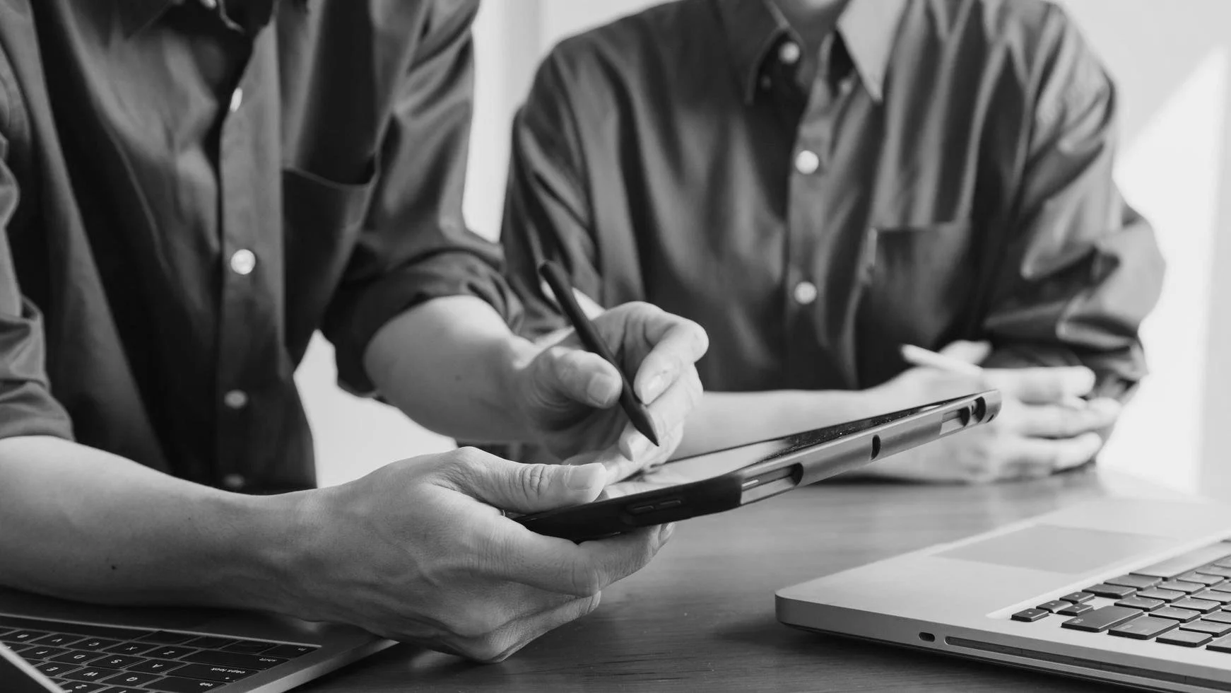 Two people in button-up shirts using a stylus and a tablet, with a laptop nearby, in a collaborative work setting.