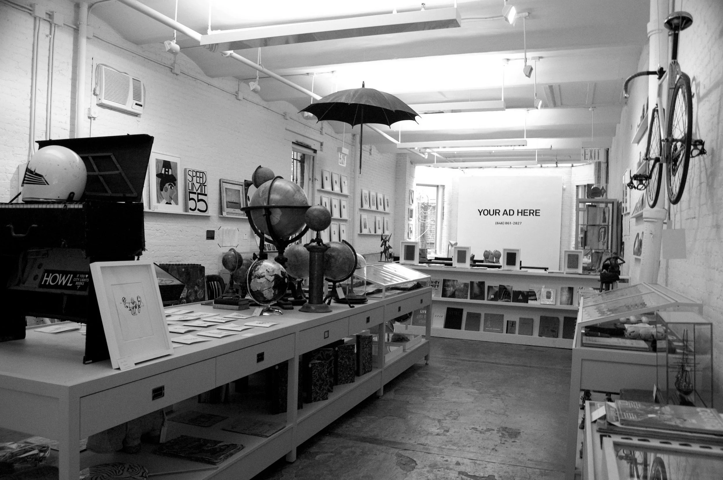Interior of an art gallery or store with display tables, globes, framed pictures on the wall, a bicycle on the wall, and a sign that says "YOUR AD HERE" in the background.