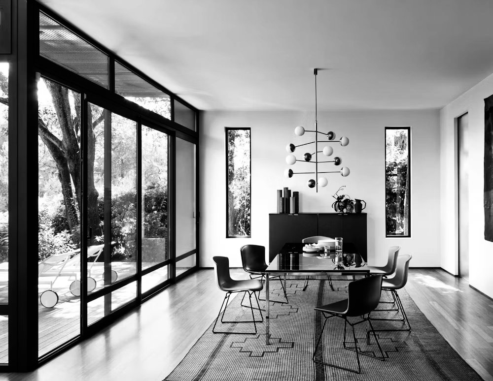 A modern dining room with a glass wall and sliding doors on the left, a rectangular glass table with six chairs, a black sideboard against the back wall, a contemporary chandelier hanging from the ceiling, and minimalist decor including books and sculptures.