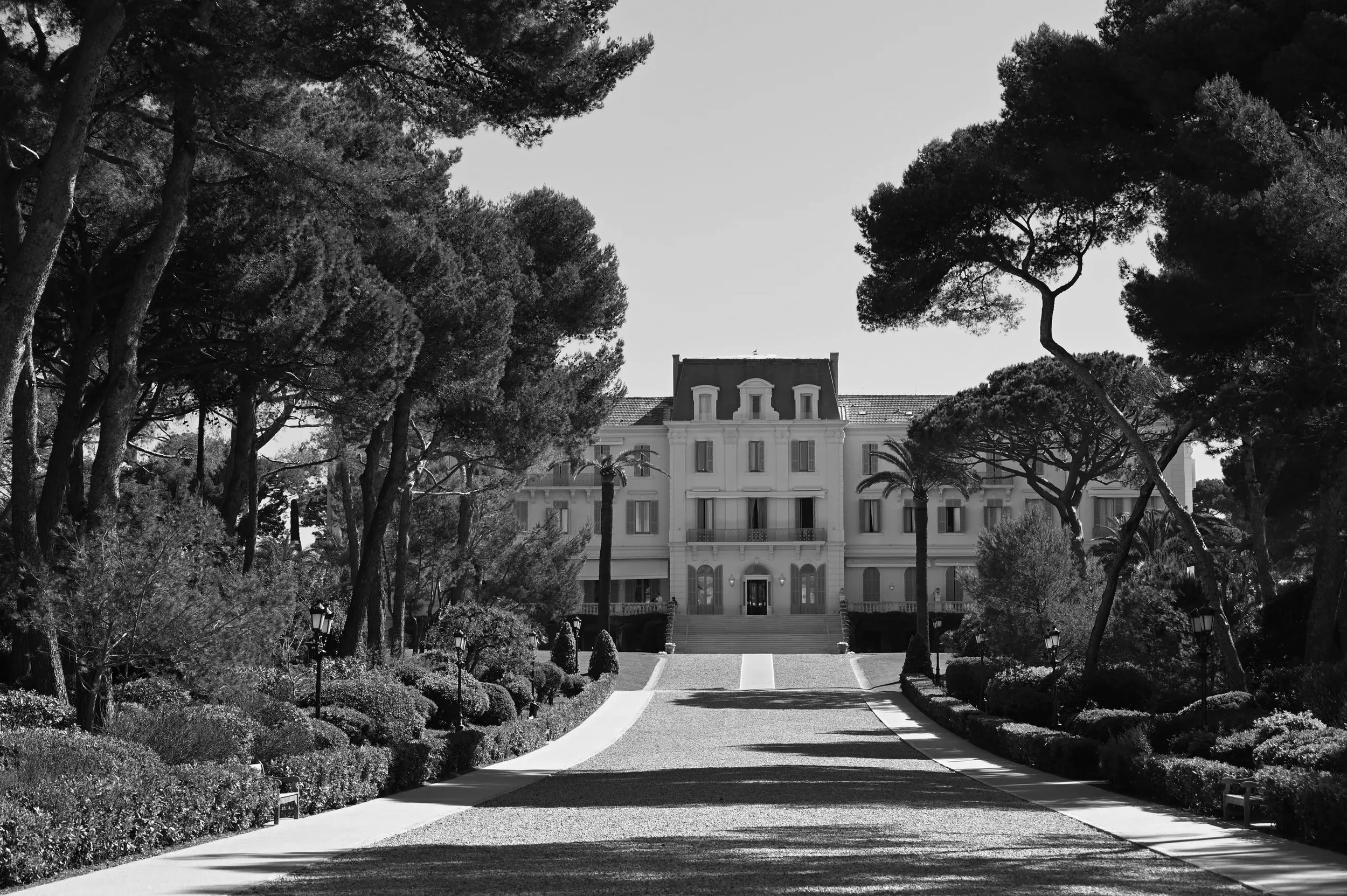 A grand, multi-story mansion with a symmetrical facade, large windows, and decorative balconies, surrounded by tall trees and well-manicured bushes, with a wide driveway leading up to the entrance.