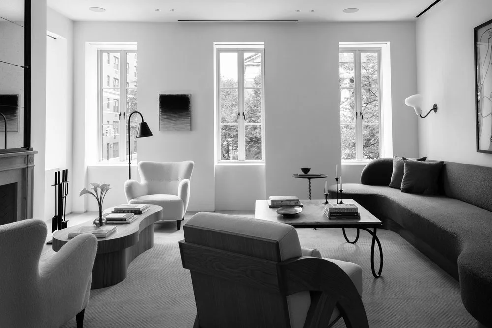 A modern living room with white walls, three large windows, a gray sectional sofa, a white armchair, a coffee table with books and decorative items, a side table with a bowl and candles, and wall art.