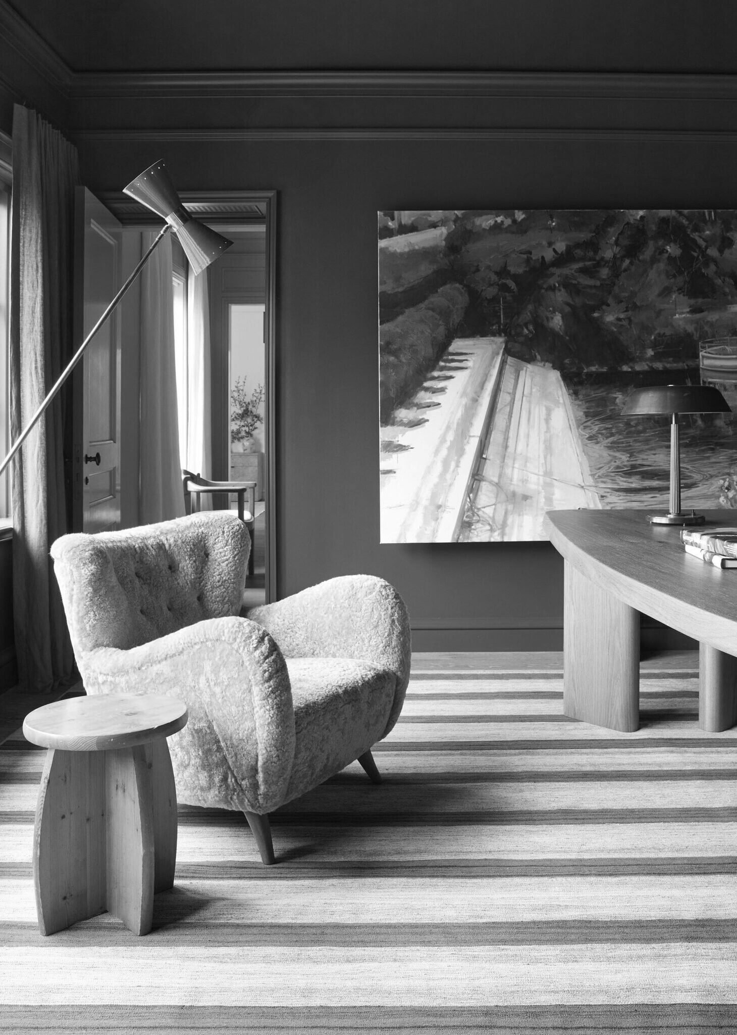 A cozy living room with a plush armchair, a small wooden side table, a curved wooden coffee table, a modern floor lamp, and a large painting on the wall. Natural light streams in through a window with curtains.