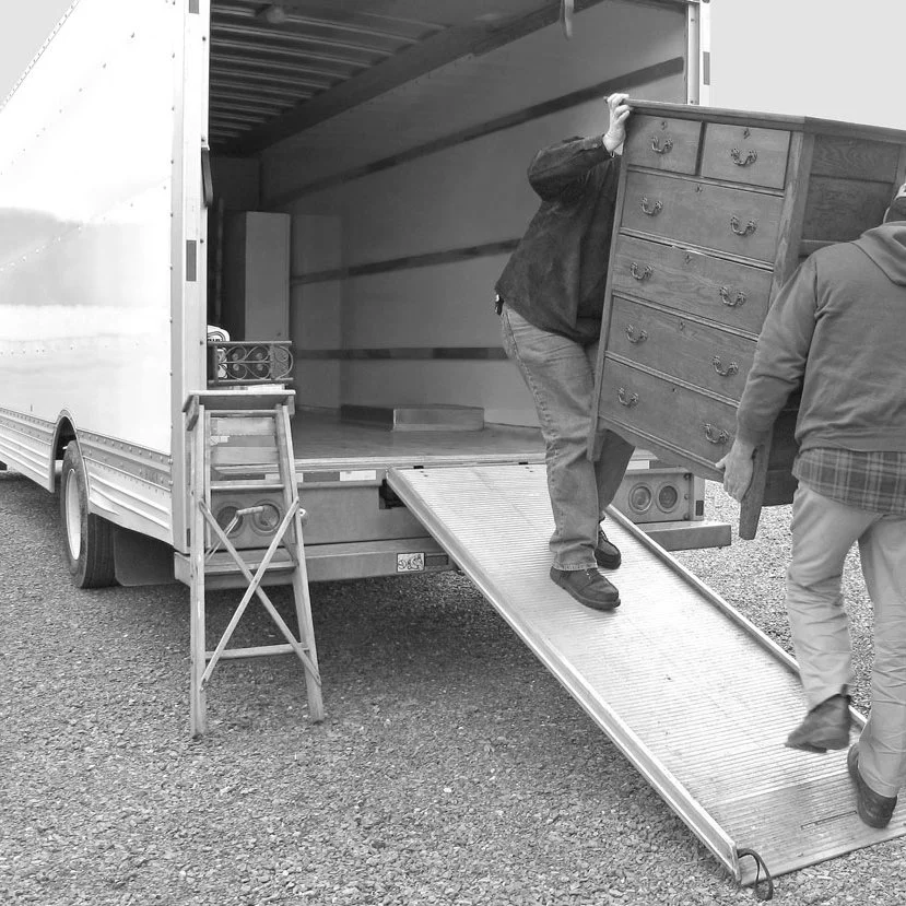 Two people unloading a wooden dresser from a moving truck using a ramp, with a small step ladder nearby.