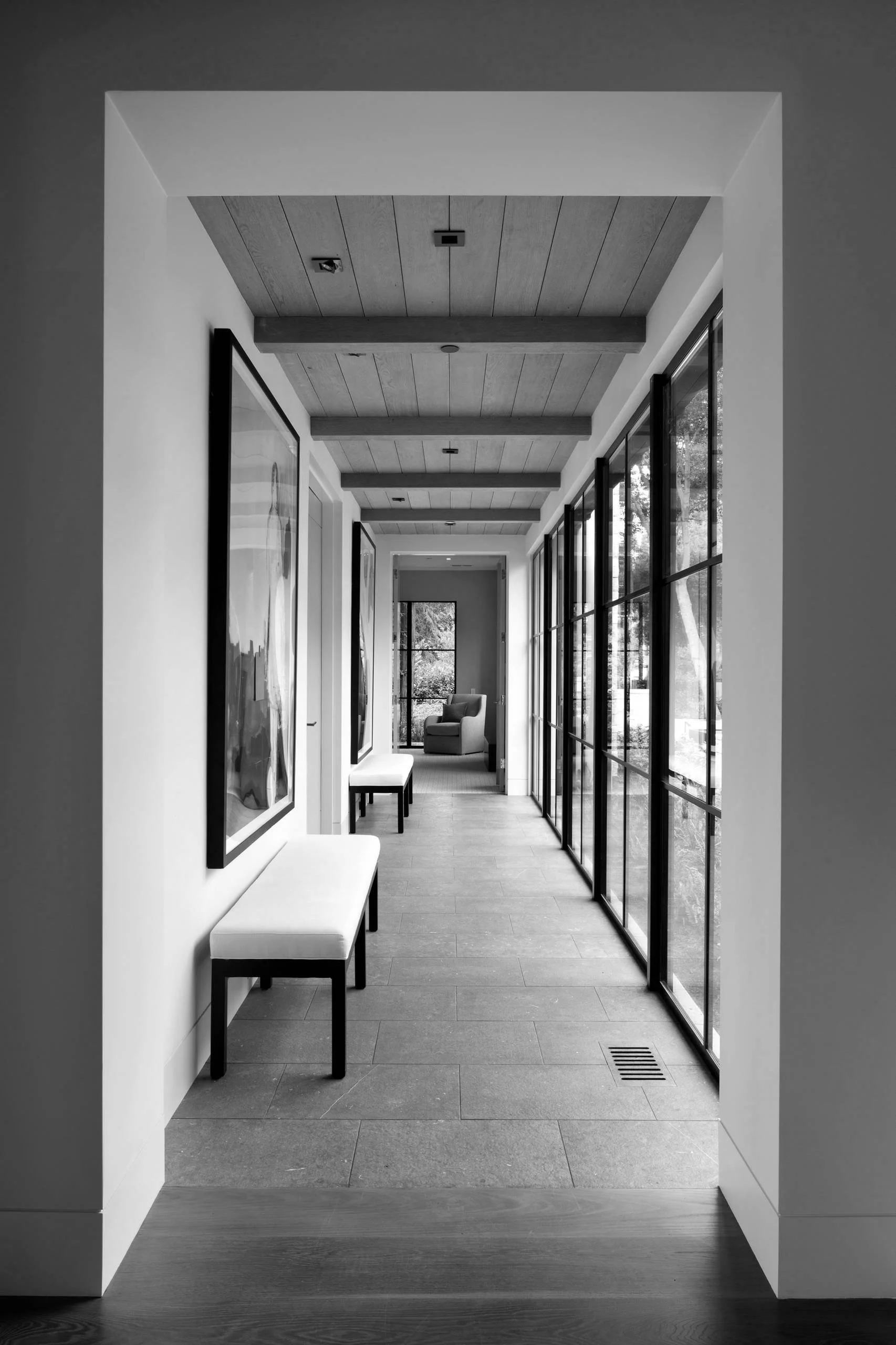 A modern, minimalist hallway with large windows on the right, framed artwork on the left, and benches along the wall, leading to a cozy corner with an armchair.