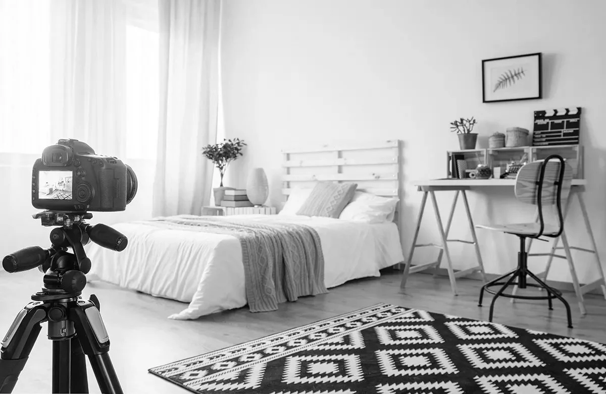 A bedroom with a bed, a desk, a chair, and a camera on a tripod, all in black and white.