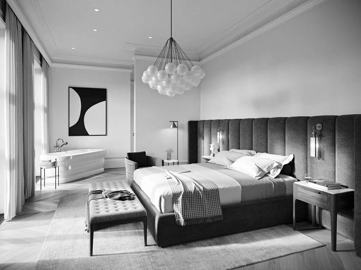 A modern hotel bedroom with a large bed, a chandelier overhead, a small table next to the bed, an armchair, a bench at the foot of the bed, a bathtub, and large windows with curtains.