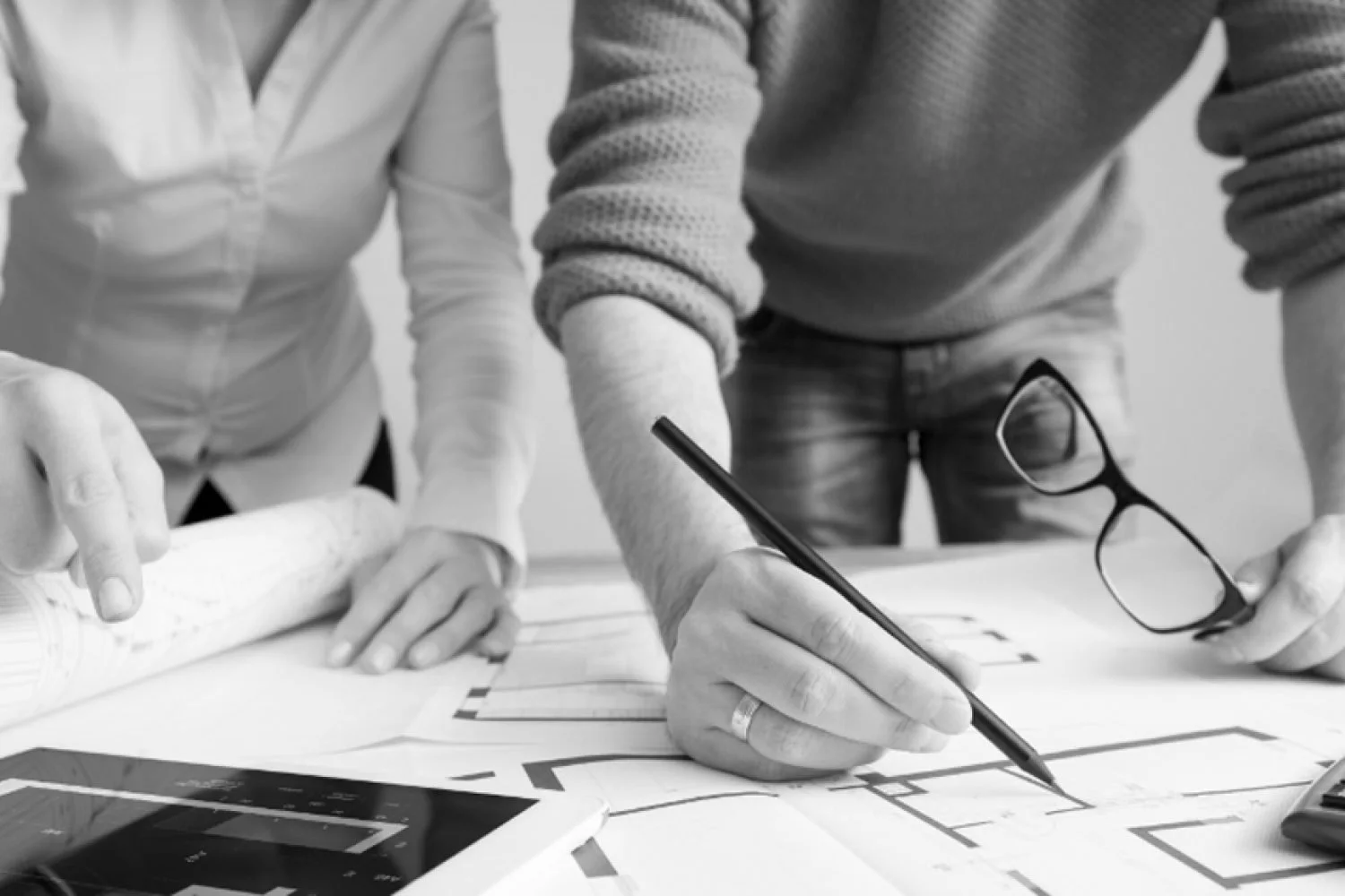 Two people are examining Blueprints on a table. One person is pointing at the blueprint with a pencil and wearing a ring, while the other holds a pair of glasses.