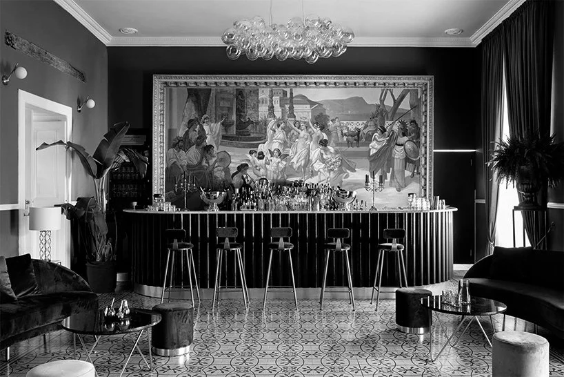 Black and white photo of a decorated interior with a bar, high chairs, a large mural of a classical scene, potted plants, curtains, sofas, and a chandelier.