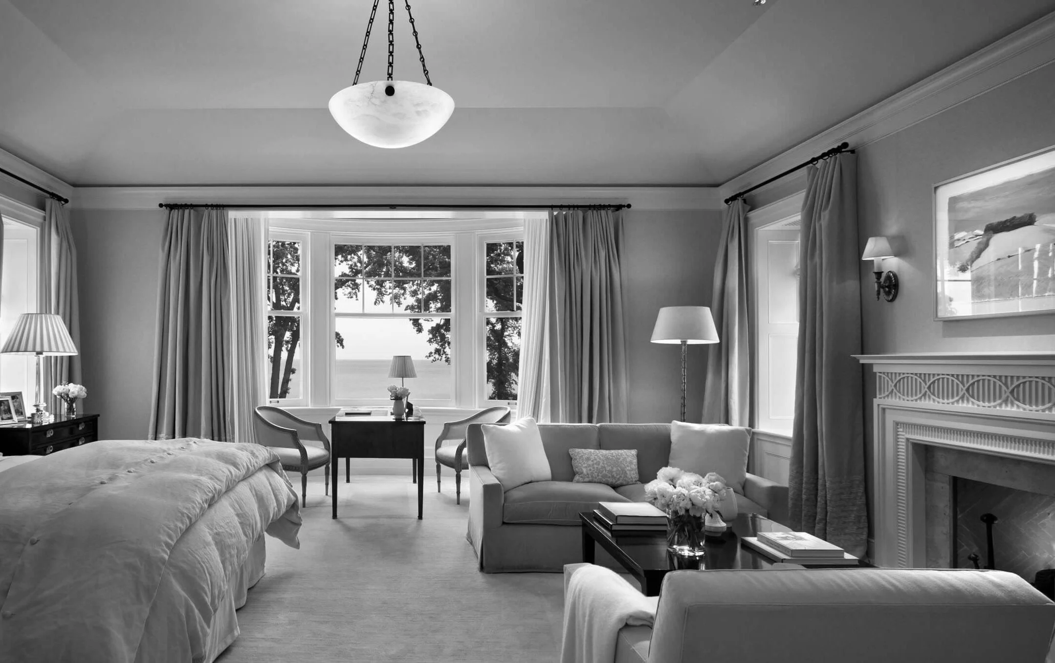Black and white photo of a living room with a large bay window showing trees outside. The room has a bed, a sofa with cushions, a coffee table with flowers, a side table with framed photos, and a fireplace.