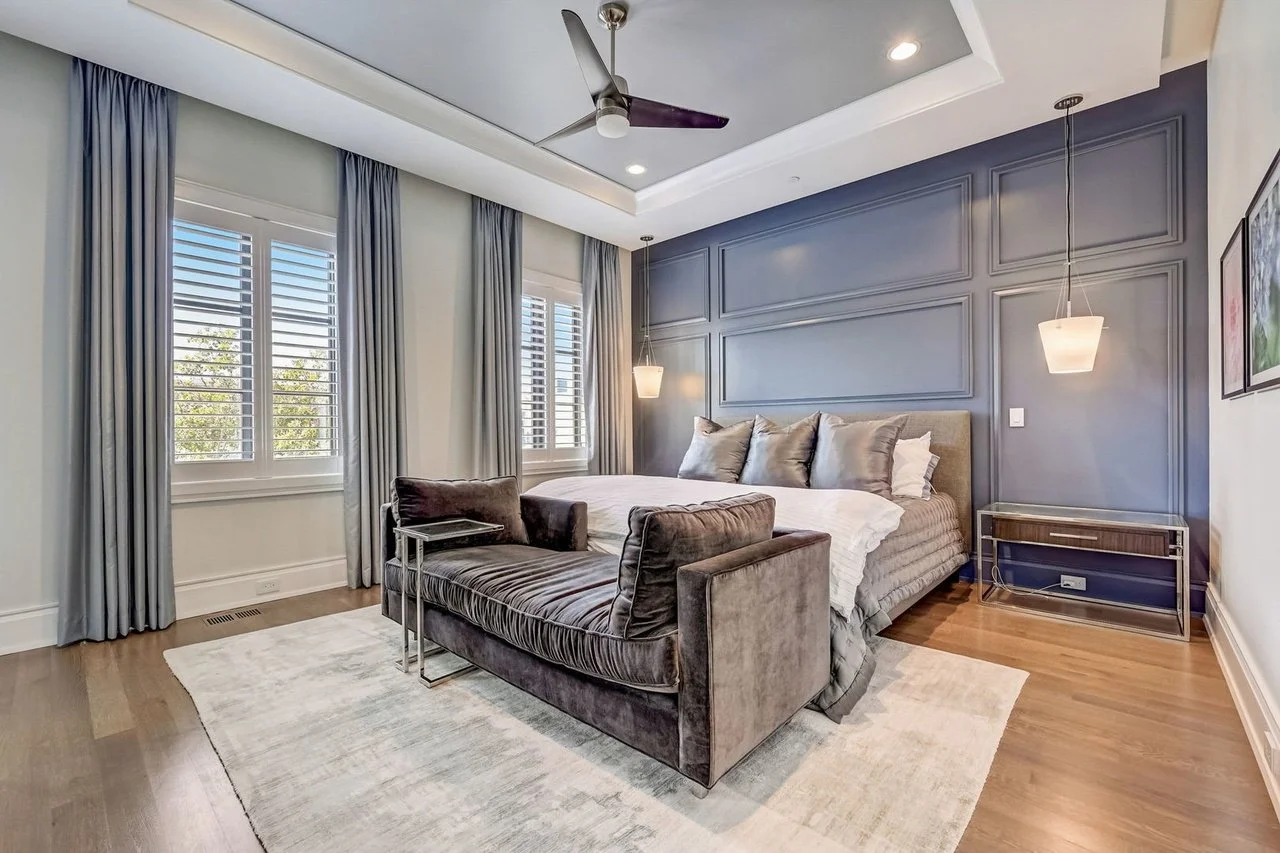 Modern bedroom with blue accent wall, large bed with silver pillows, dark gray velvet chaise lounge, two windows with white blinds and light gray curtains, hardwood floors, wall-mounted pendant lights, ceiling fan, framed art on wall.