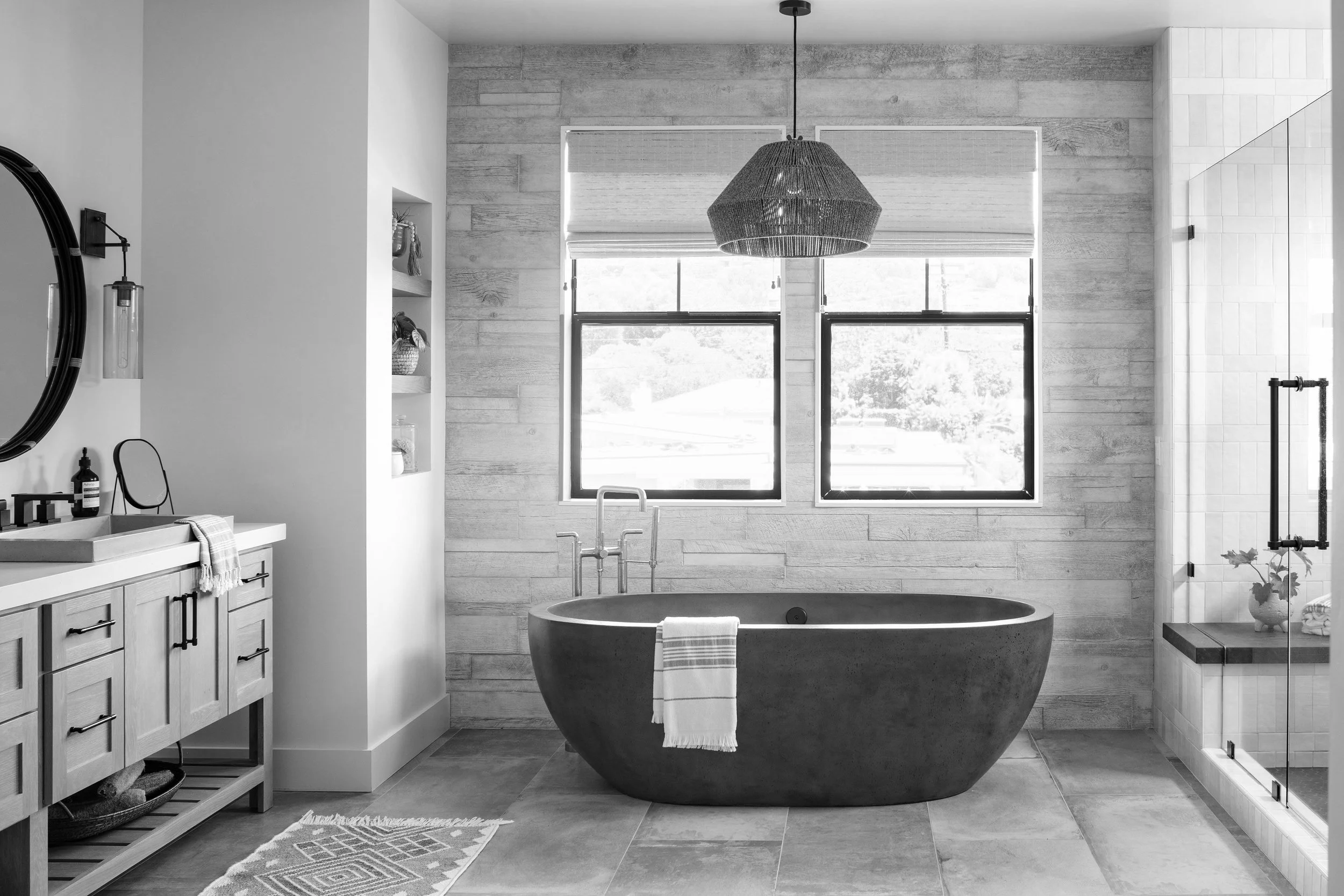 A modern bathroom with a freestanding bathtub under two large windows, a vanity with a mirror and sink on the left, and a walk-in shower with glass door on the right, all in black and white.