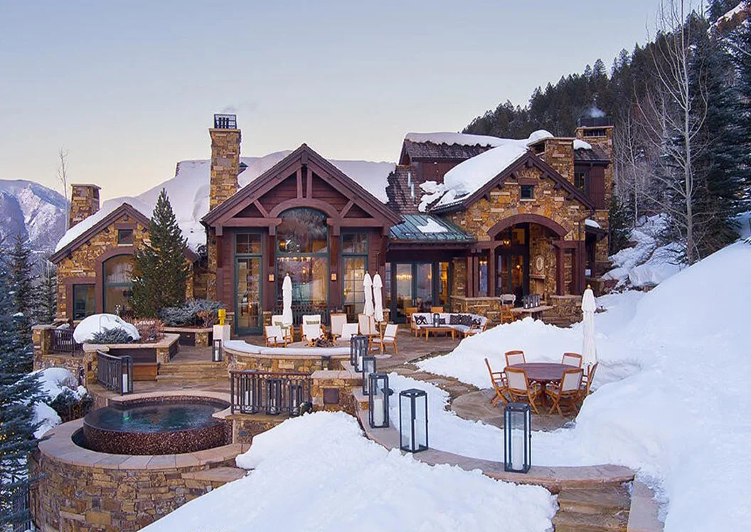 A large mountain house with stone and wood exterior, surrounded by snow-covered landscape, outdoor seating, umbrellas, and a hot tub.