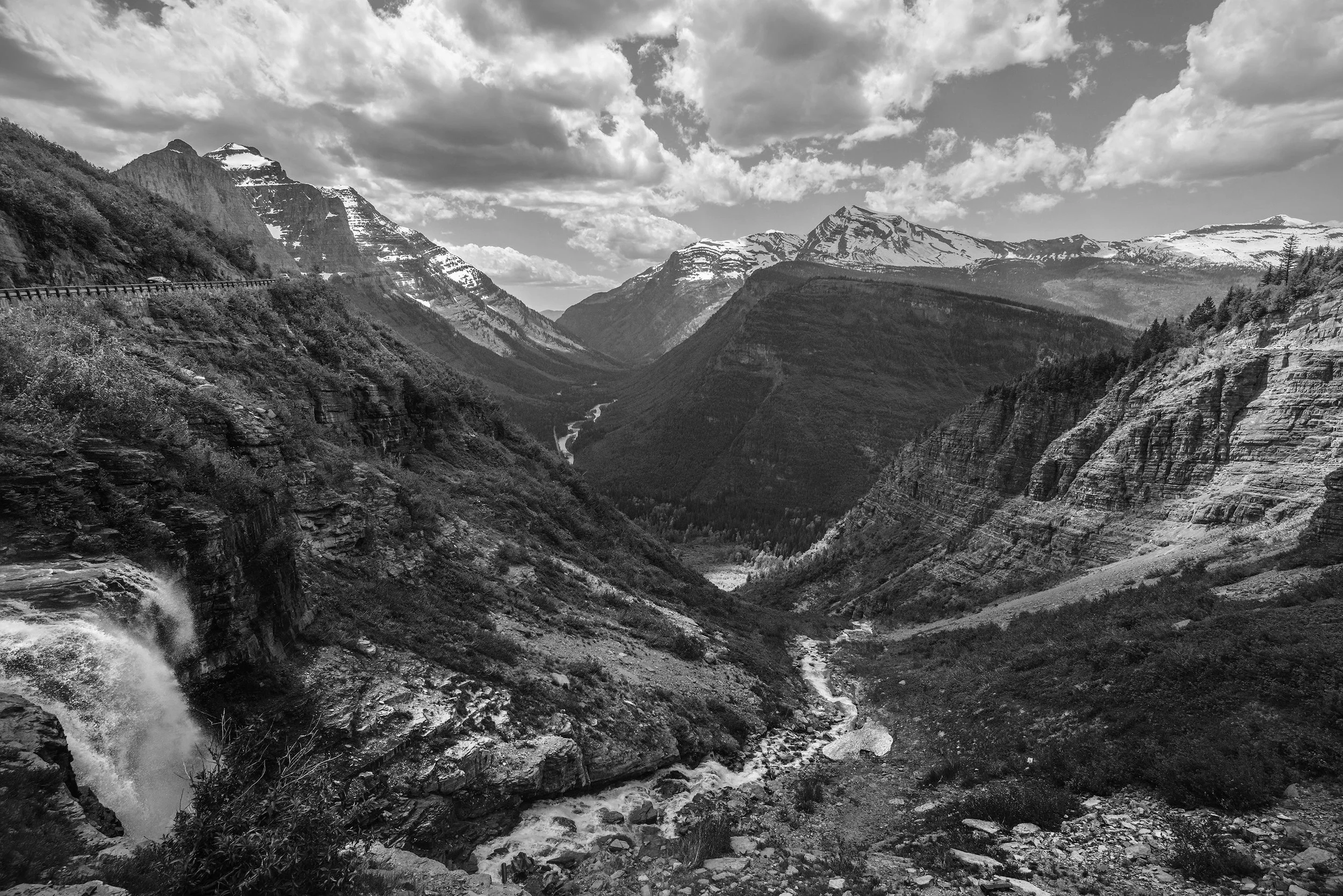 Black and white photo of a mountainous canyon with snow-capped peaks, a river flowing through the valley, and a waterfall on the left.